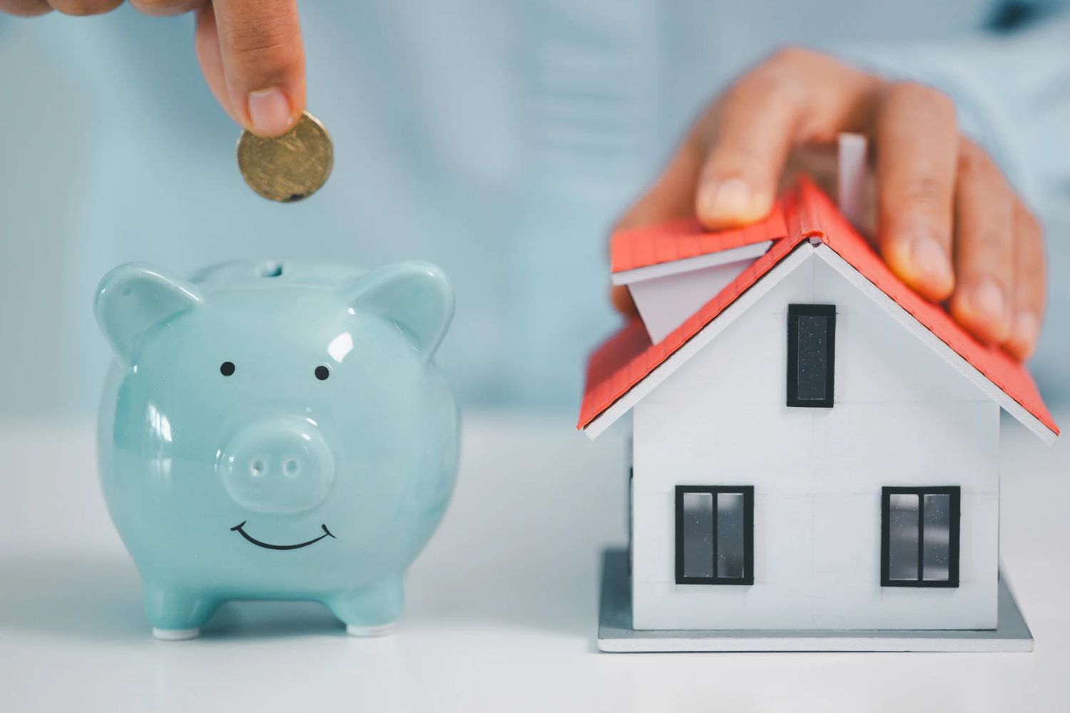 Person placing a coin into a piggy bank next to a model house, representing home equity and savings.